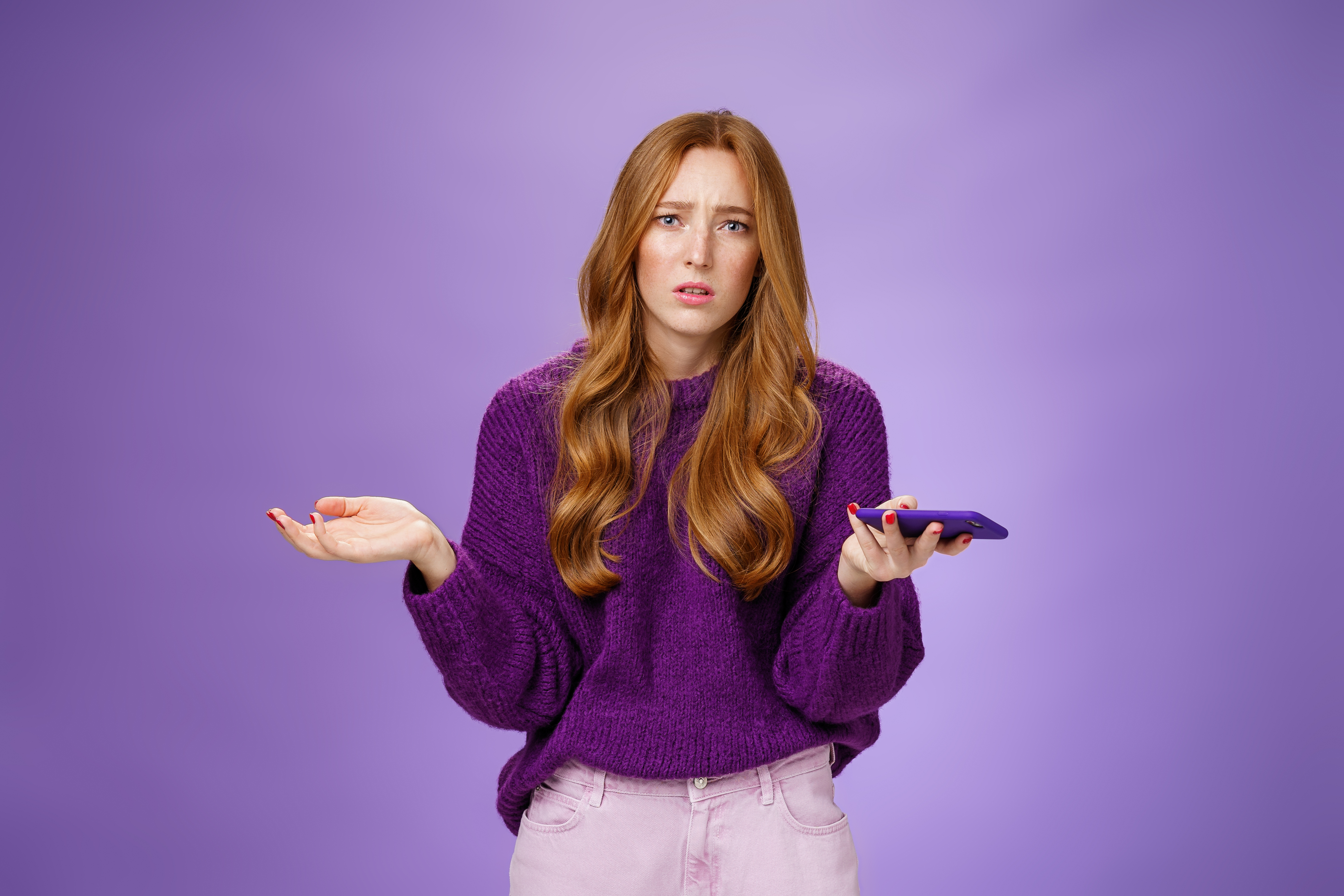 Clueless and sad cute redhead woman spread hands sideways frowning disturbed and upset holding smartphone being questioned and clueless why mobile phone not working, posing over purple wall